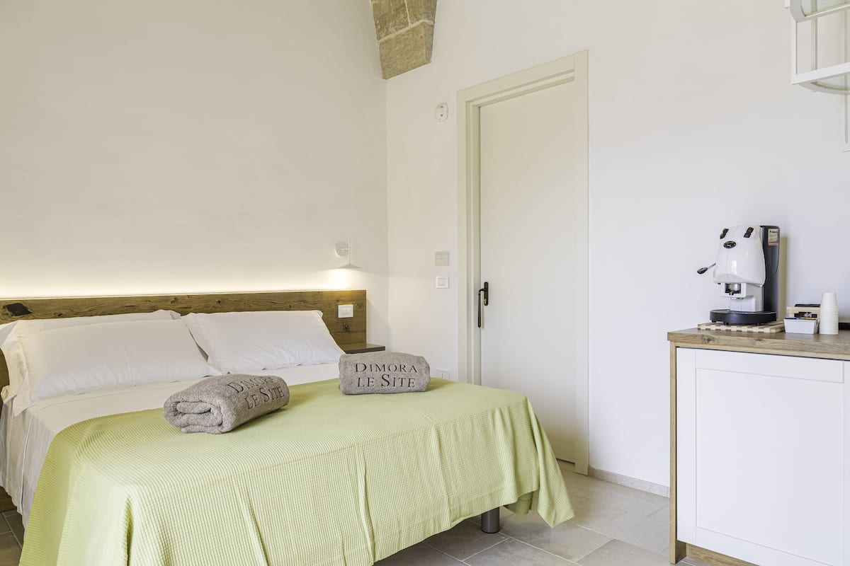 A neatly made bed with a green bedspread, a pillow with "Dimora Le Site" text, and a stone with the same text.
