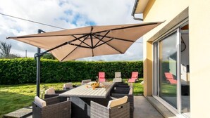 En udendørs terrasse med et bord og stole, en stor parasol og en glasdør, der fører til en have.