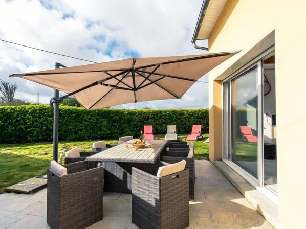 An outdoor patio with a table and chairs, a large umbrella, and a glass door leading to a garden.