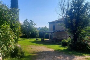 A rustic stone building with a covered porch, surrounded by lush greenery and a clear sky.
