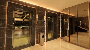 Un ascenseur moderne doté de portes en verre, une poubelle et un distributeur de gel hydroalcoolique, le tout situé dans un hall d’entrée aux murs de marbre et agrémenté d’un escalier.