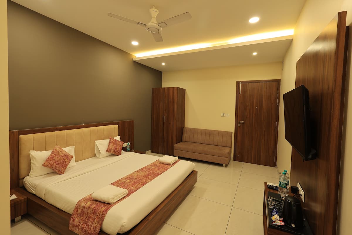 A modern bedroom with a large bed, a sofa, a wardrobe, a television, and a desk with a water bottle and remote controls.