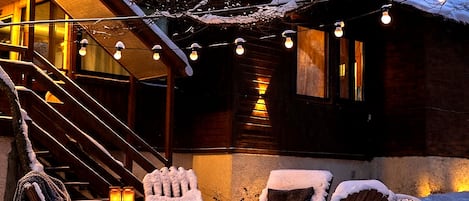 Un área de estar al aire libre cubierta de nieve, con un brasero, sillas de madera y una escalera que lleva a un edificio.