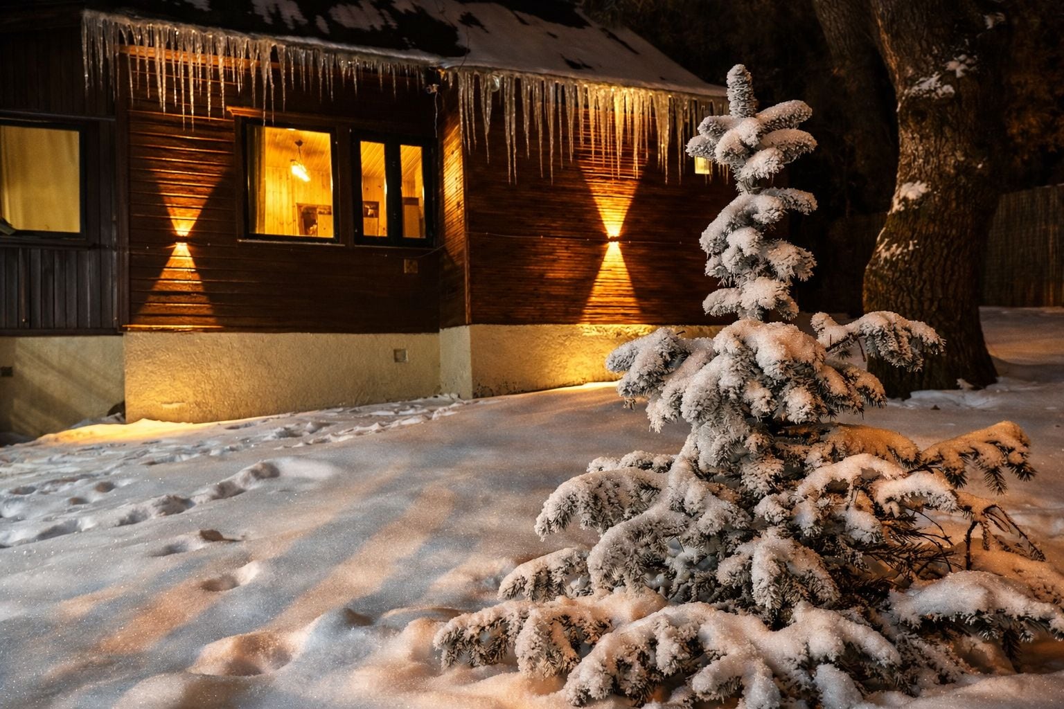 A cabin with a snow-covered ground, icicles hanging from the roof, and a snow-covered tree in front.