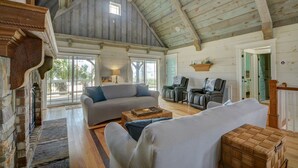 A spacious living room with a high ceiling, wooden beams, a grey sofa, a white armchair, and a wooden coffee table.