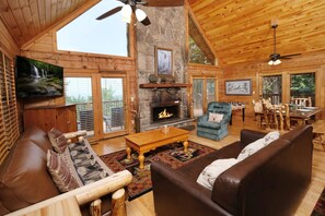 Interior acogedor de una cabaña con chimenea de piedra, muebles de madera y ventanas amplias con vistas al exterior.