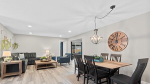 Un salon moderne avec un coin repas, comprenant une grande horloge en bois, un lustre et un ensemble de canapés.