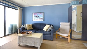 A living room with a blue sofa, a white side table, and a chair. There is a painting on the wall and a window with curtains.