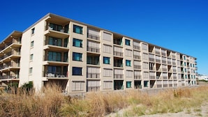 A multi-story hotel with balconies, located near a beach with dry grass and sand.