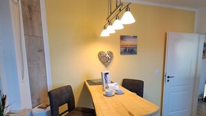 Uma área de refeições com uma mesa de madeira, duas cadeiras, um lustre e uma porta branca ao fundo.