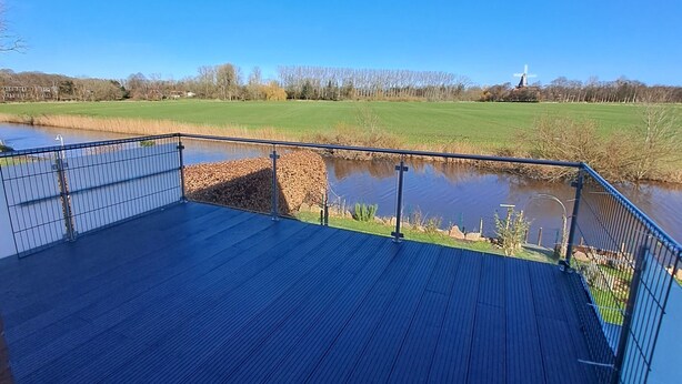 Um deck de madeira com guarda-corpo de metal, vista para um rio e um moinho de vento ao longe.