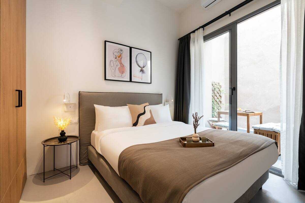 A modern hotel room with a large bed, a bedside table with a lamp, and a view of the outdoors through a sliding door.