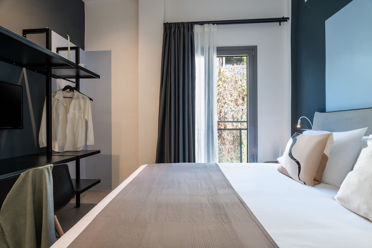 A modern hotel room with a large bed, a black metal shelf unit, a hanging chair, and a view of a tree through a window with curtains.