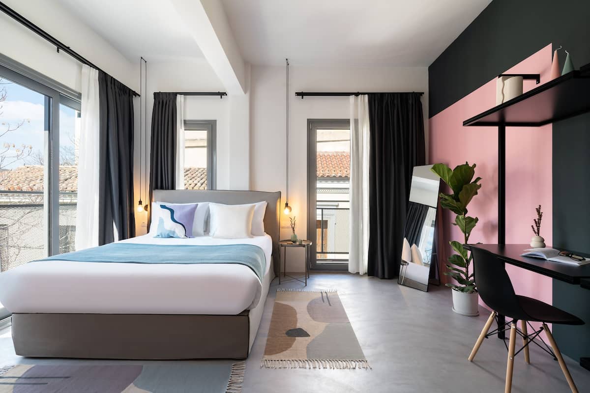 A modern hotel room with a large bed, a desk, a chair, a potted plant, and a view of the outside.
