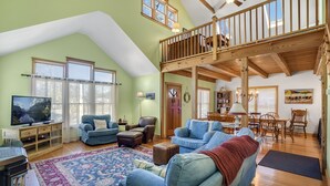 A spacious living room with a high ceiling, wooden beams, a balcony, and a large TV. - Family Tides-311 139th St-Bayside W/D, AC, WIFI (Ocean City)