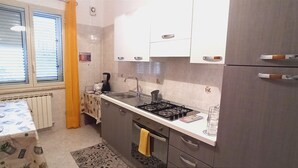 A compact kitchen with a gas stove, sink, and a refrigerator. There is a window with blinds and a curtain, and a patterned tablecloth on the table. - Vacation home byron (Contessa Entellina)