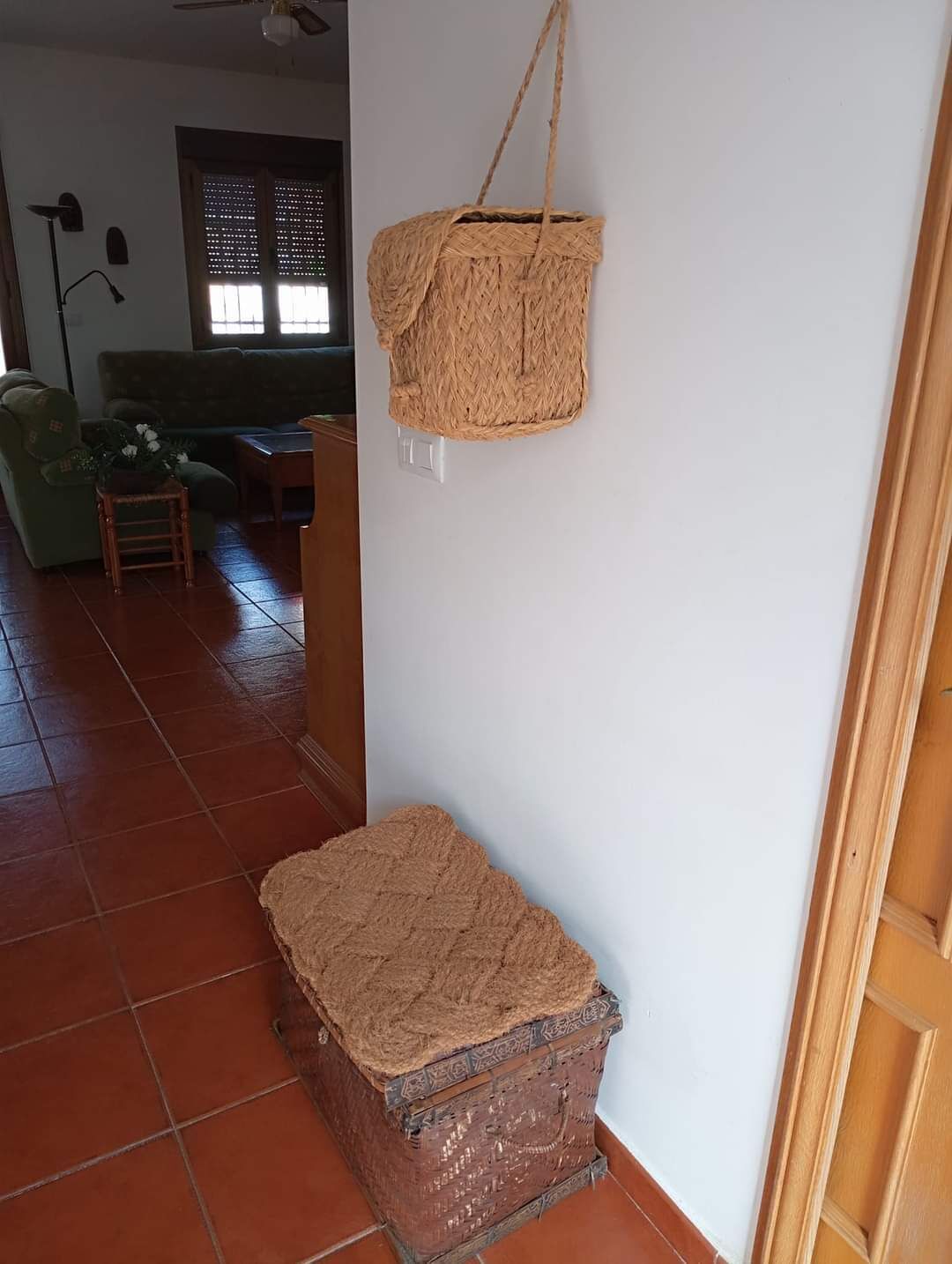 Una habitación con una cesta tejida colgada en la pared, un pequeño taburete con una planta y un aparador tejido junto a la pared.