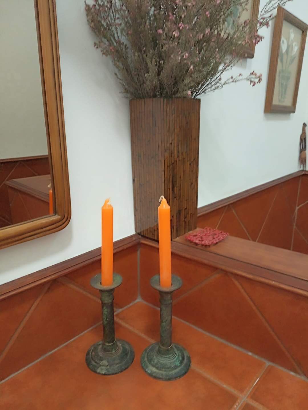 Deux bougies posées sur un sol carrelé, un pot de fleurs en bois, un miroir et une petite statue.