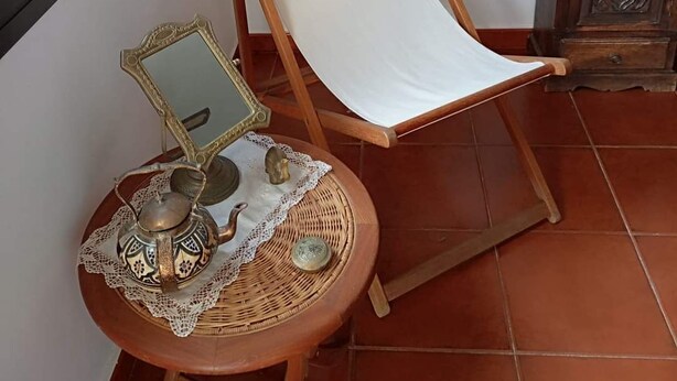 A room with a wooden chair with a white canvas backrest, a small round table with a decorative item, a lamp, and a cabinet.