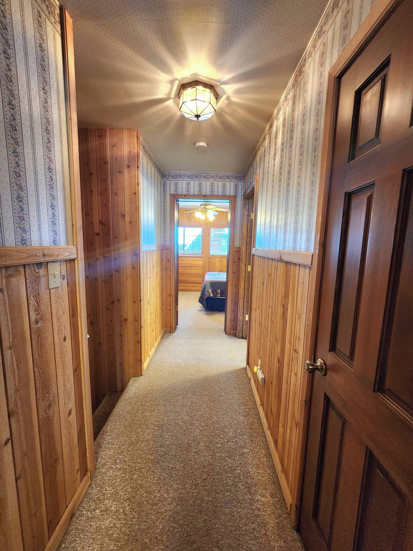 A hallway with wooden walls and a carpeted floor leading to a room with a bed and a ceiling fan.