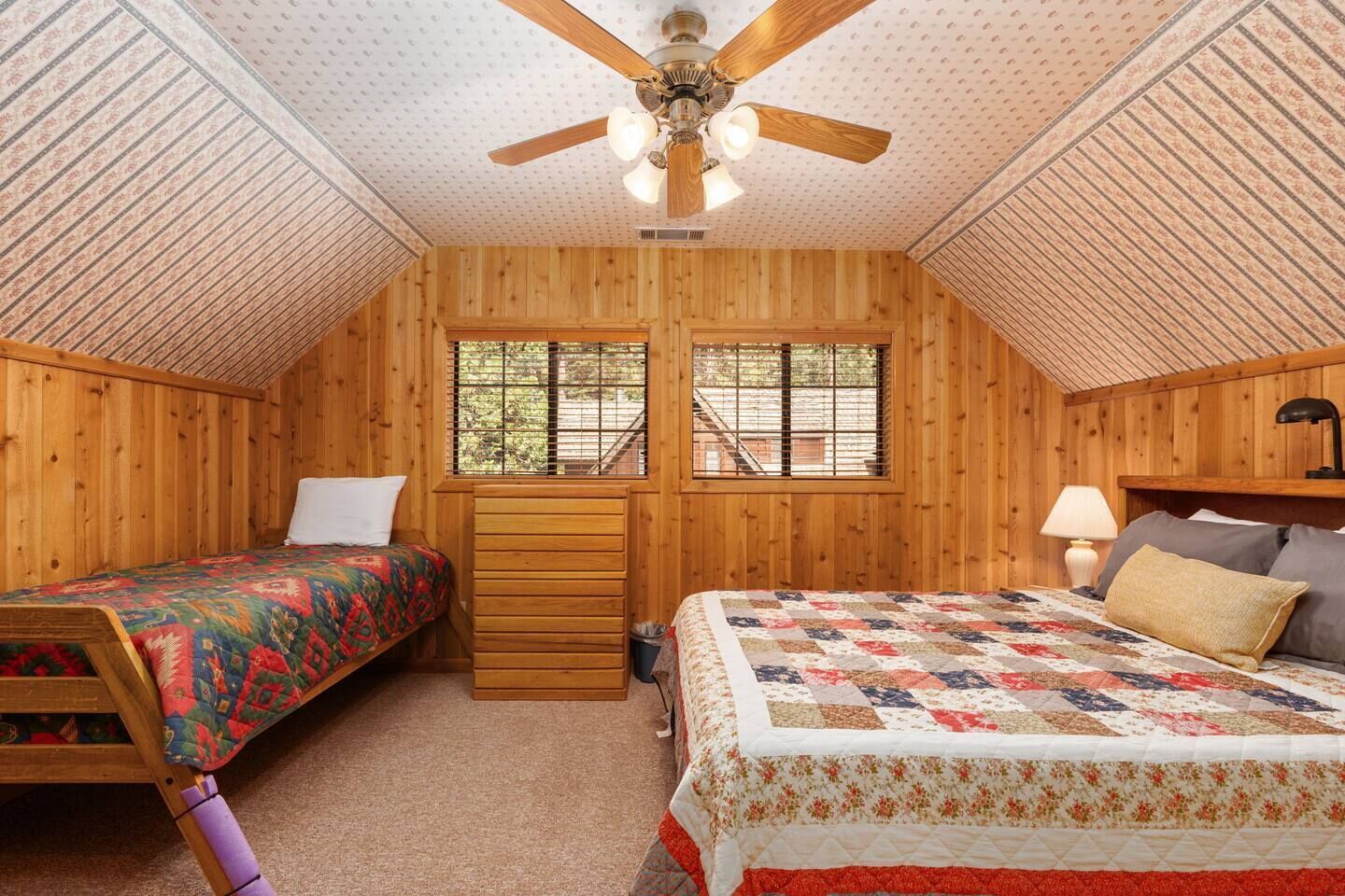 Un dormitorio acogedor en el ático con paredes de madera, un ventilador de techo, dos camas con colchas de colores, un aparador y una ventana con vista a una escalera.