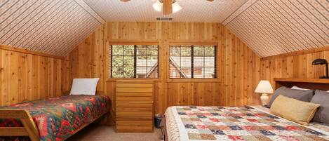 Un dormitorio acogedor en el ático con paredes de madera, un ventilador de techo, dos camas con colchas de colores, un aparador y una ventana con vista a una escalera.