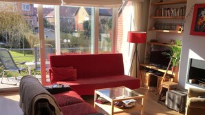 Un salón con un sofá rojo, una mesa de centro y un mueble de televisión con televisor de pantalla plana. Hay una estantería con libros y objetos decorativos, y un ventanal que ofrece vistas al exterior.