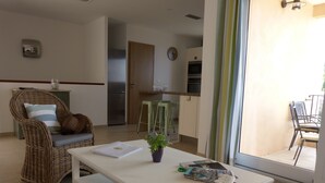 Uma sala de estar com um sofá de vime, uma mesa de centro branca com revistas e vista para uma varanda com cadeiras.