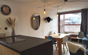 A modern kitchen with a central island, dining table, and chairs. There is a decorative mirror, a wall-mounted light fixture, and a window with a view of the outside.