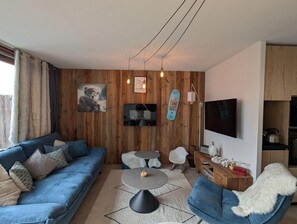 A modern living room with a blue sofa, two round tables, a TV mounted on the wall, and wooden paneling.