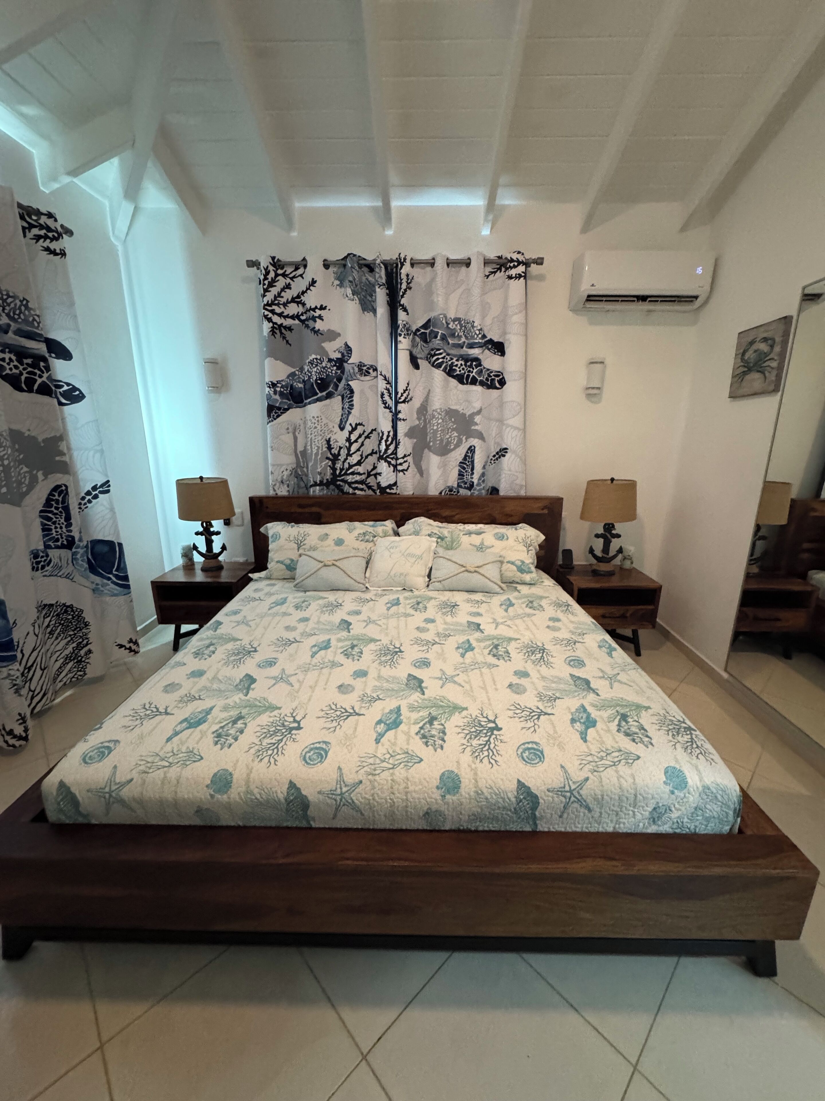 A bedroom with a wooden bed, two bedside tables with lamps, a window with curtains, and an air conditioning unit on the wall.