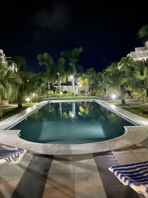 A nighttime swimming pool with surrounding palm trees and a building in the background.