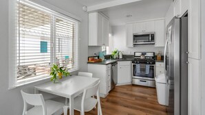 A modern kitchen with white cabinets, a stainless steel oven, a microwave, and a refrigerator. There is a dining table with chairs and a window with blinds. - OCEAN VIEW|Steps to the Beach, Main St. & Pac City (Huntington Beach)