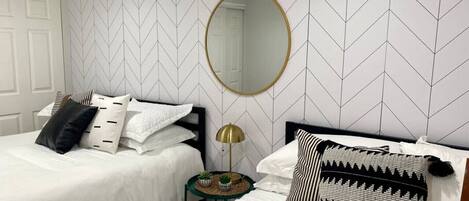A hotel room with two beds, a round mirror, a small table with a lamp, and a patterned throw pillow.