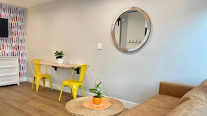 A living room with a brown sofa, yellow chairs, a round table with a plant, a round mirror, and a TV on the wall. - Steps To Beach, Main St, Pac City | BBQ | Firepit (Huntington Beach)