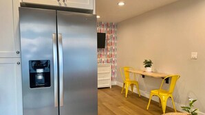 A compact kitchen with a stainless steel refrigerator, a small dining area with a wooden table and two yellow chairs, and a wall-mounted TV. - Steps To Beach, Main St, Pac City | BBQ | Firepit (Huntington Beach)