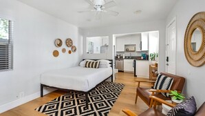 Een moderne, goed verlichte kamer met een bed, een stoel, een klein tafeltje en een keuken in de achtergrond.