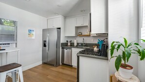 Een moderne keuken met witte kasten, een roestvrijstalen koelkast, een vaatwasser en een gasfornuis. Er is een klein eiland met een waterkoker en een bloempot.