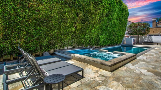 Una piscina con solárium de piedra, rodeada por un seto verde exuberante y un patio con sillones de descanso.