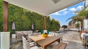 Un patio exterior con mesa y sillas de madera, rodeado de piso de piedra y un seto verde exuberante.