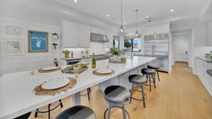 A modern kitchen with white cabinetry, a long island with bar stools, stainless steel appliances, and pendant lighting. - Rooftop Ocean Views / 2min Walk To Beach / Private Pool (Huntington Beach)
