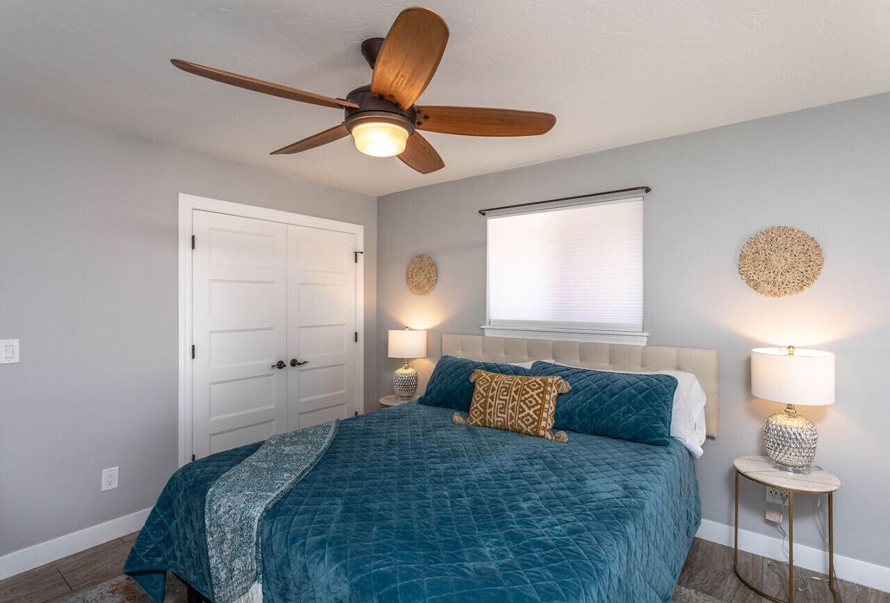 A bedroom with a bed, ceiling fan, bedside lamps, and a window with blinds.