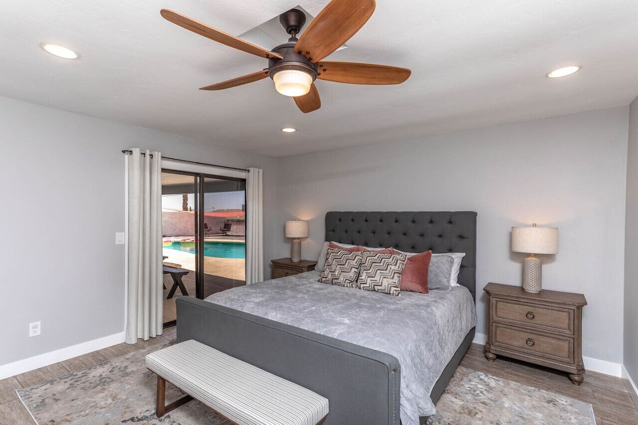 A bedroom with a bed, a bench, a ceiling fan, two bedside tables with lamps, and a view of a swimming pool through a glass door.