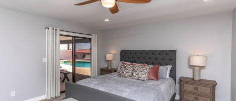 A bedroom with a bed, a bench, a ceiling fan, two bedside tables with lamps, and a view of a swimming pool through a glass door.
