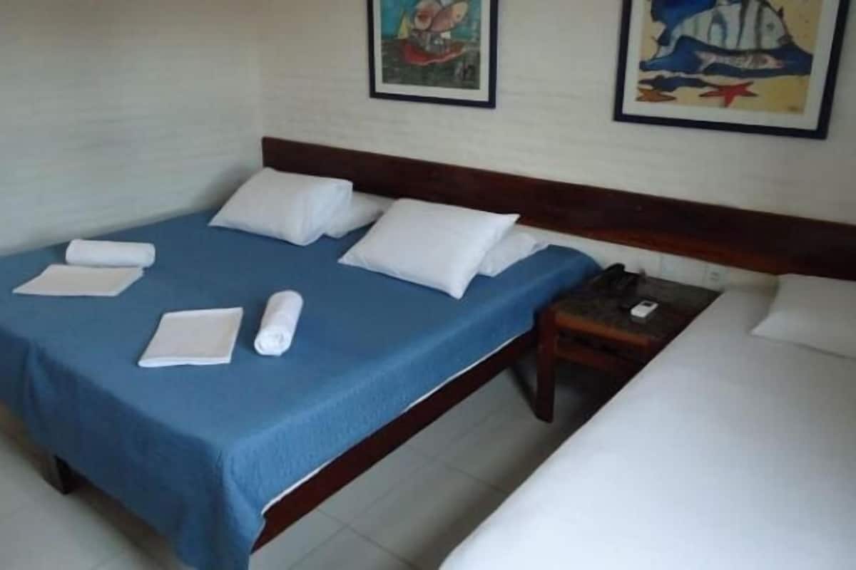 A hotel room with a bed, pillows, a blue bedspread, a wooden headboard, a nightstand, a phone, and two paintings on the wall.