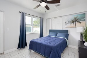 Ein Schlafzimmer mit Bett, Deckenventilator, Fenster mit Vorhängen, einem Wandbild und einem Nachttisch mit Lampe.