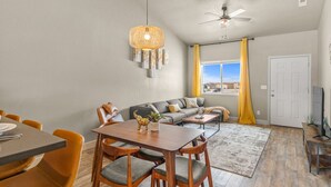 Una sala moderna con comedor, un conjunto de sofás, un ventilador de techo y un ventanal con cortinas.