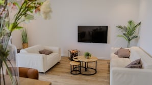 Een moderne woonkamer met twee witte banken, een salontafel, een aan de muur bevestigde televisie en een potplant.
