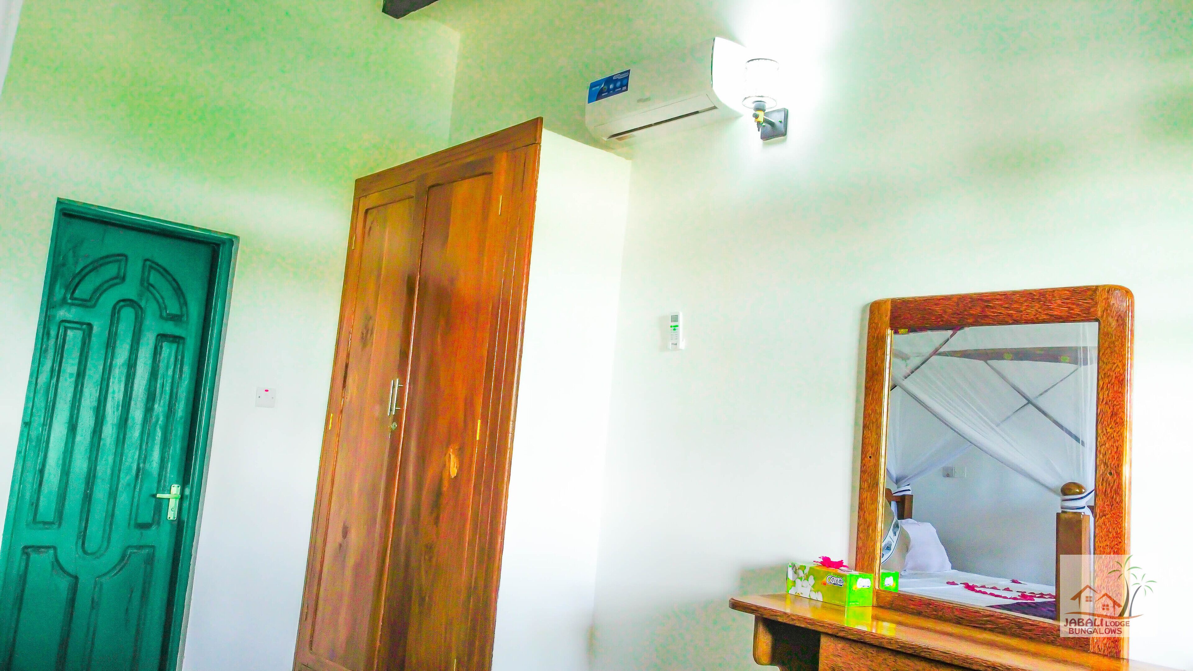 A room with a green door, a wooden wardrobe, a mirror with a wooden frame, and a small table with a tissue box.
