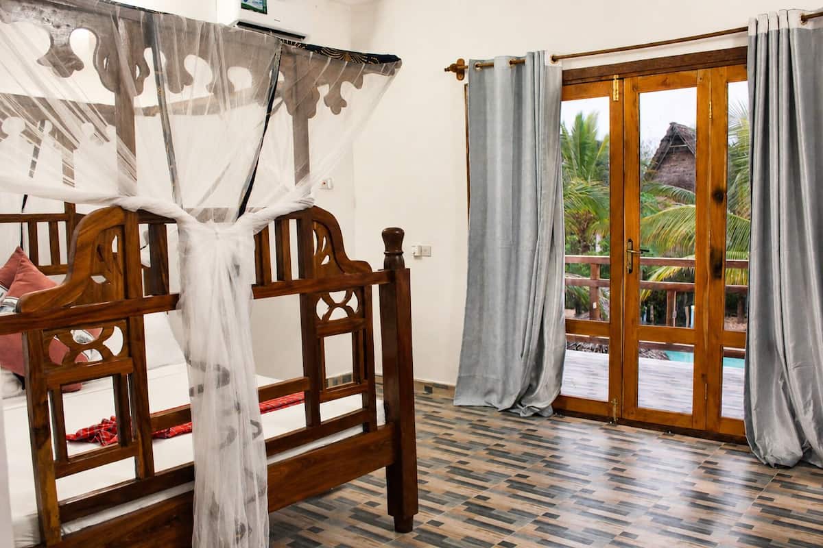 A room with a wooden bed, a window with curtains, and a view of palm trees outside.